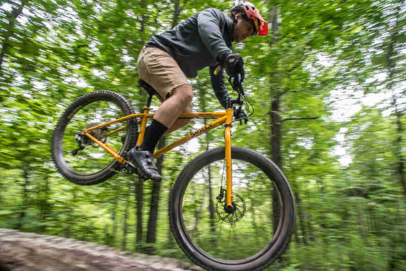 Right side view of a cyclist going airborne on a yellow Surly bike, with trees in the background 