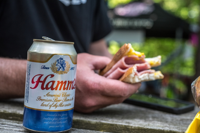 A can of Hamm's beer in front of a person's hand holding a ham sandwich, at a picnic table