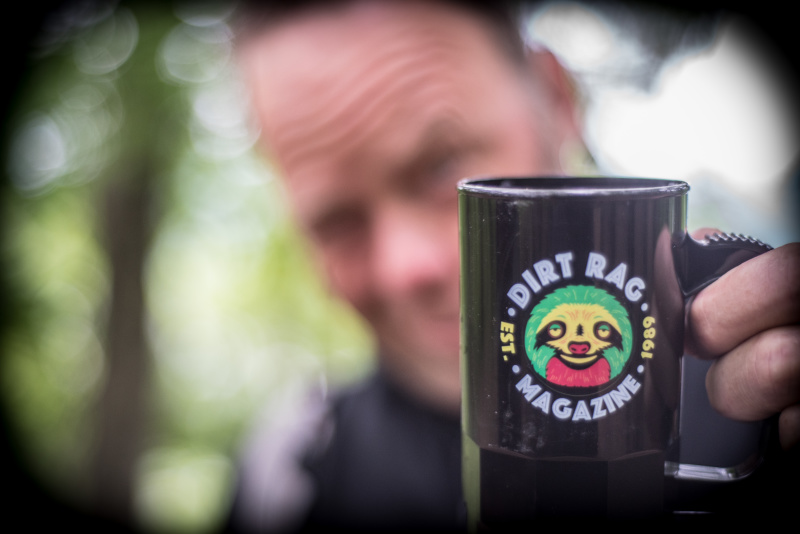 A black coffee mug with, Dirt Rag, shown on it, being held by a person blurred out in the background
