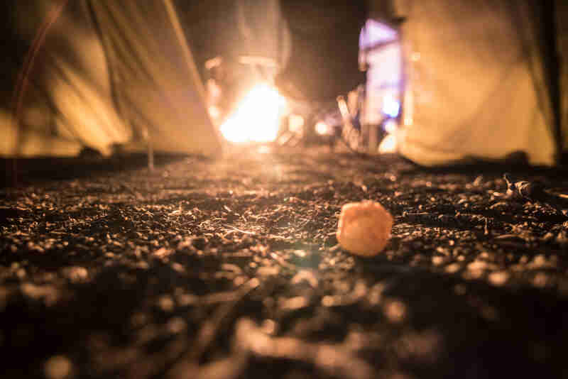 A ground level view of a cheese ball laying in dirt, with a campfire between two tents in the background, at night