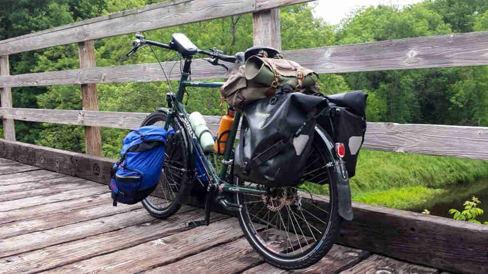 Rear, left side view of a green Surly Disc Trucker bike, parked on a wood bridge, along the handrail, with trees behind