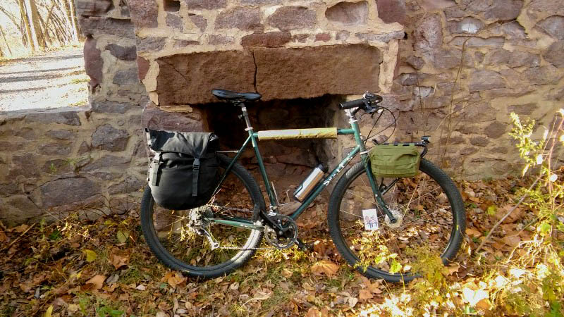 Right side view of a Surly Disc Trucker bike, green, standing in leaves in front of an outdoor fireplace