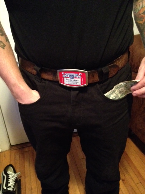 Front, chest down view of a person wearing black Surly pants and a Budweiser belt buckle, in a room with wood floors