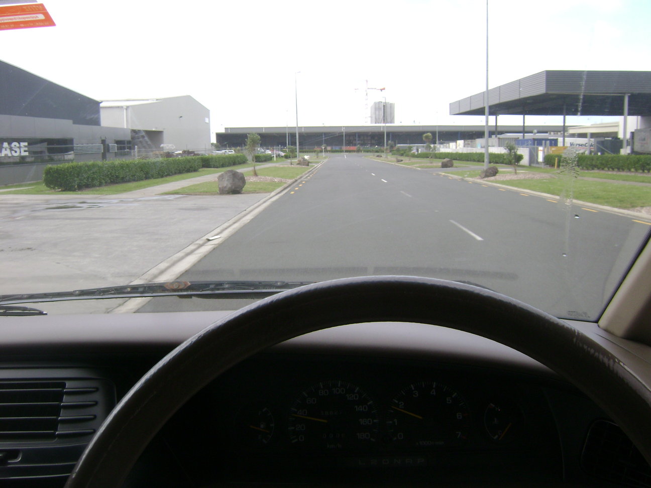 View from behind the steering wheel of a car that's facing straight away on a roadway, with warehouse buildings ahead
