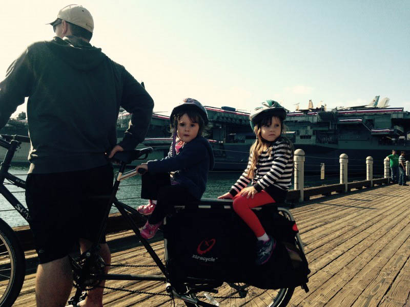 Right side view of a Surly Big Dummy bike with 2 children on back and a cyclist standing over, on a wood pier