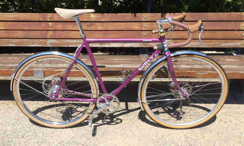 Right side view of a raspberry color Surly bike, parked on a sidewalk, alongside a park bench