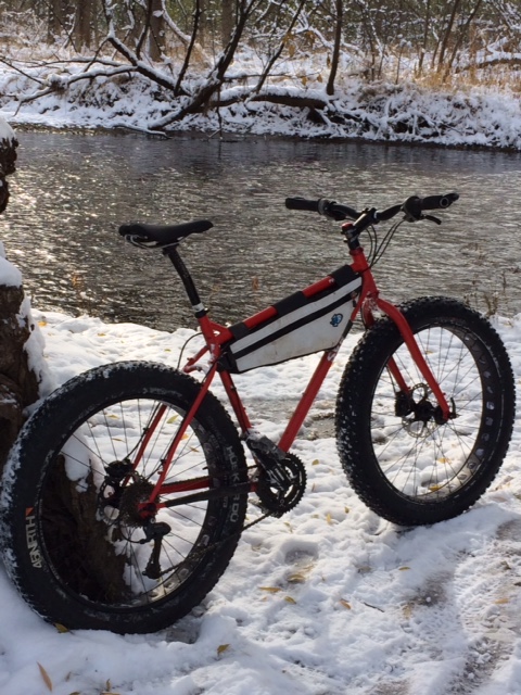 Right side view of a red Surly fat bike, parked on a snowy riverbank , with the woods in the background