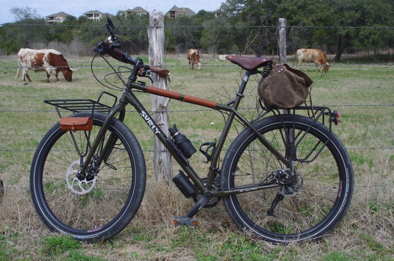 Left side view of a Surly ECR bike, leaning against a barbed wire fence post, with cows in a pasture behind it