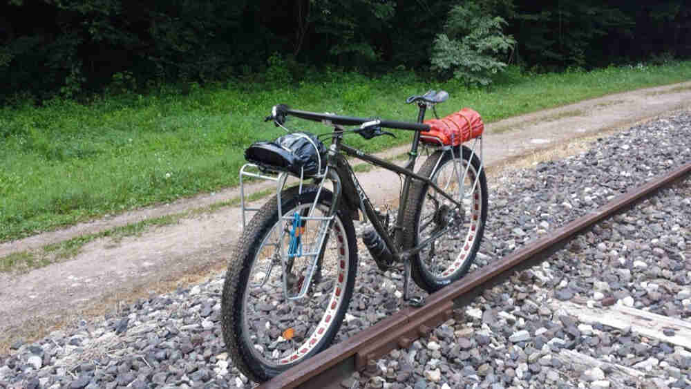 Front, right side view of a Surly ECR bike, parked along a train track rail, next to a dirt trail with trees behind