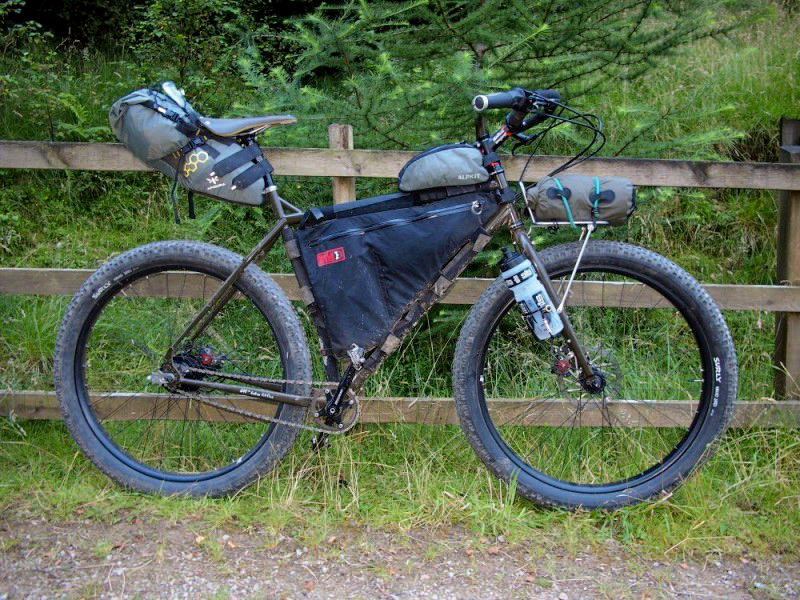 Right side view of a Surly ECR bike, olive, against a wood fence, with thick trees in the background