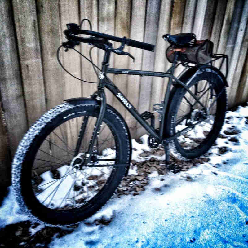 Left side view of a black Surly ECR bike, parked on snowy rocks against a wood fence wall