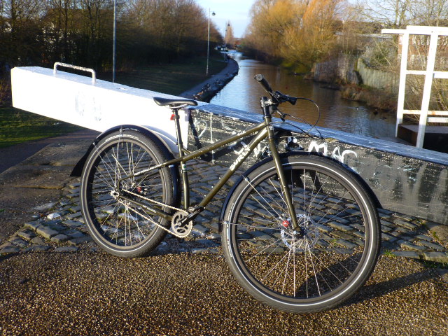Right side view of an olive drab Surly ECR bike, leaning on a railing, with a stream flowing straight away in background