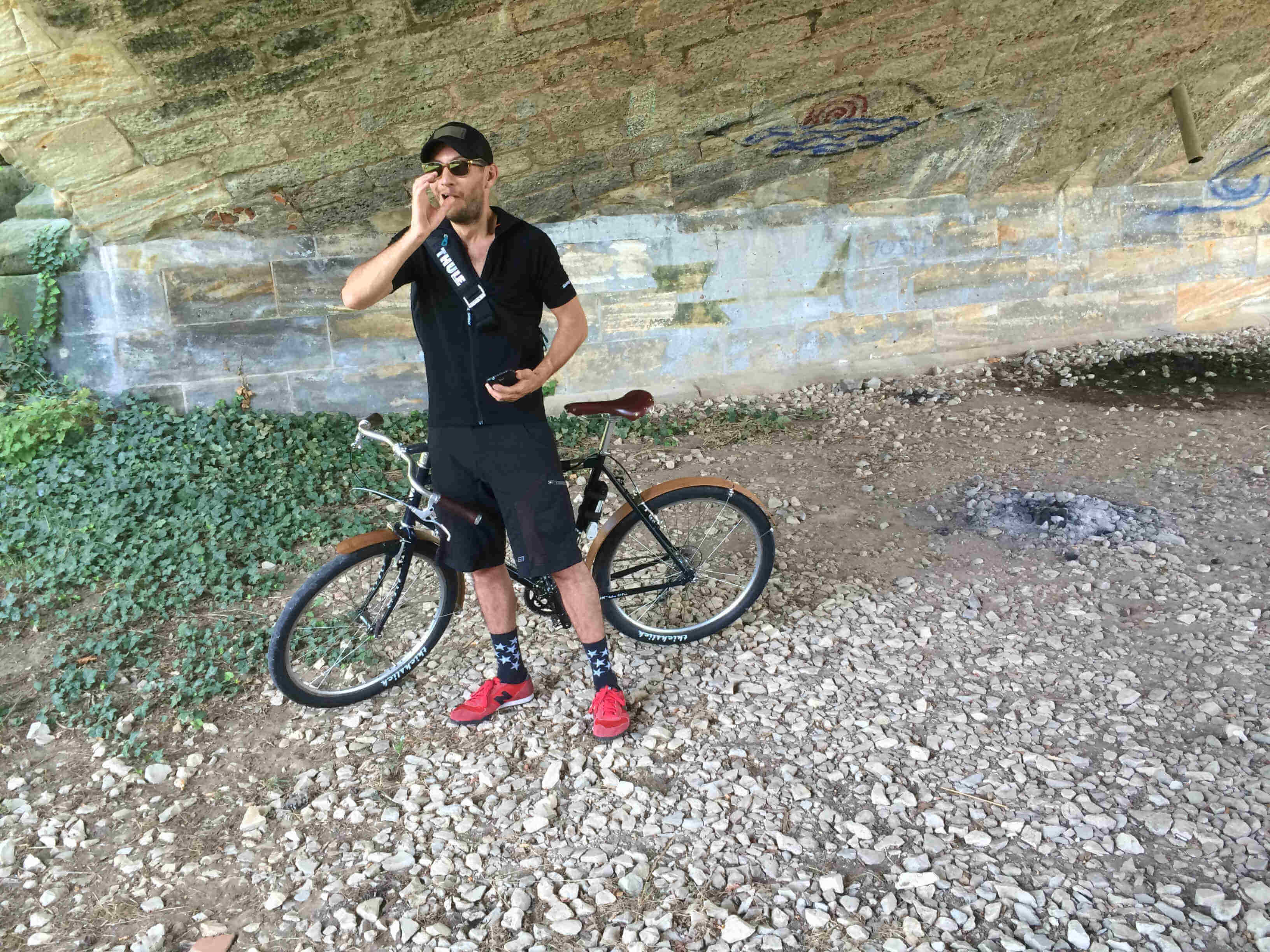 Front view of a cyclist, standing in front of the left side of a black Surly bike, on gravel under a stone overpass