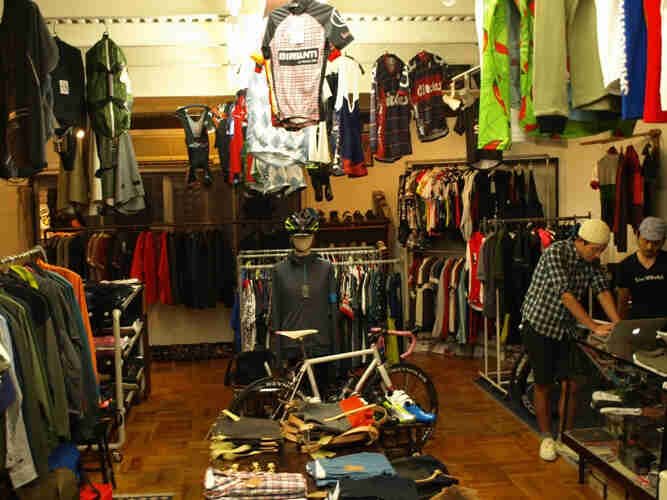 Inside view of a shop with bicyclist clothing on retail racks, with 2 people looking at a laptop, on the right
