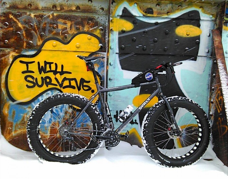 Right side view of gray Surly fat bike with a NASA top bar pack, parked on snow, in front of a wall with graffiti