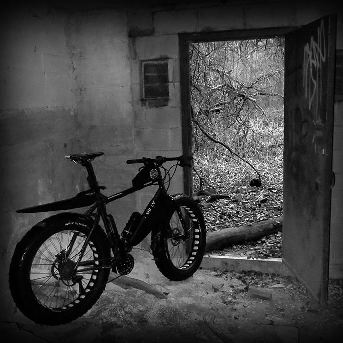 Black & white image - Rear, right side view of a Surly fat bike, parked inside a building,  in front of an open door