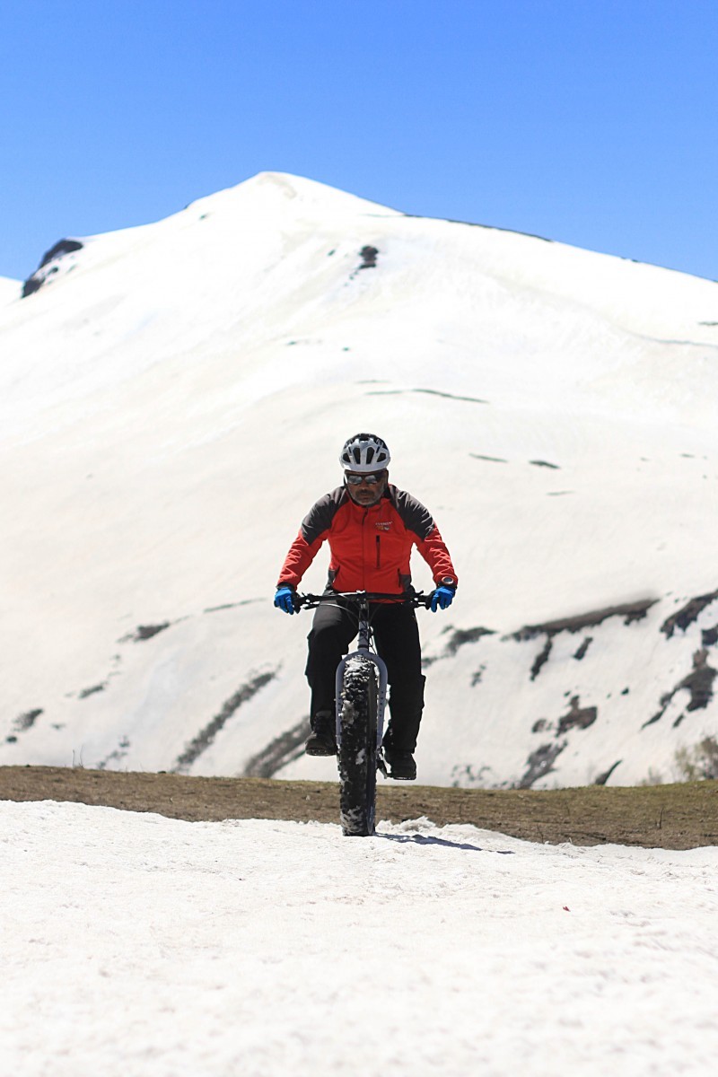 Front view of a cyclist, riding a Surly fat bike on a snowy patch, with a snow covered mountain in the background