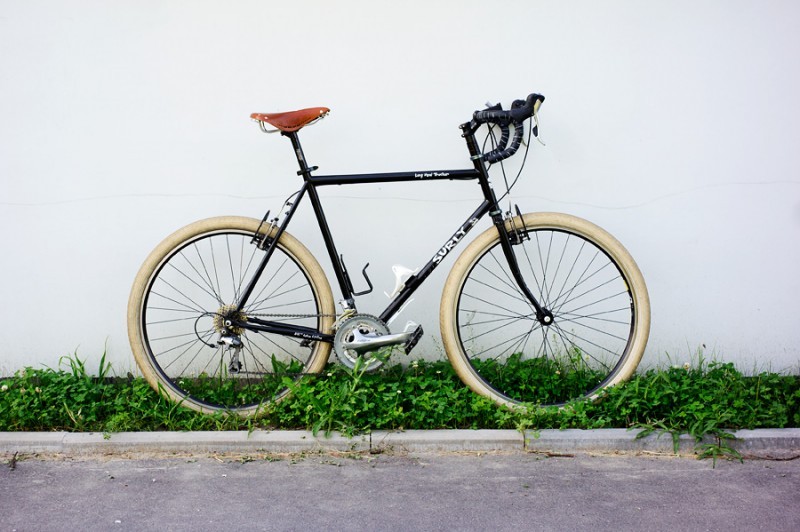Right side view of a black Surly Long Haul Trucker bike with white tires, parked on weeds, against a white wall