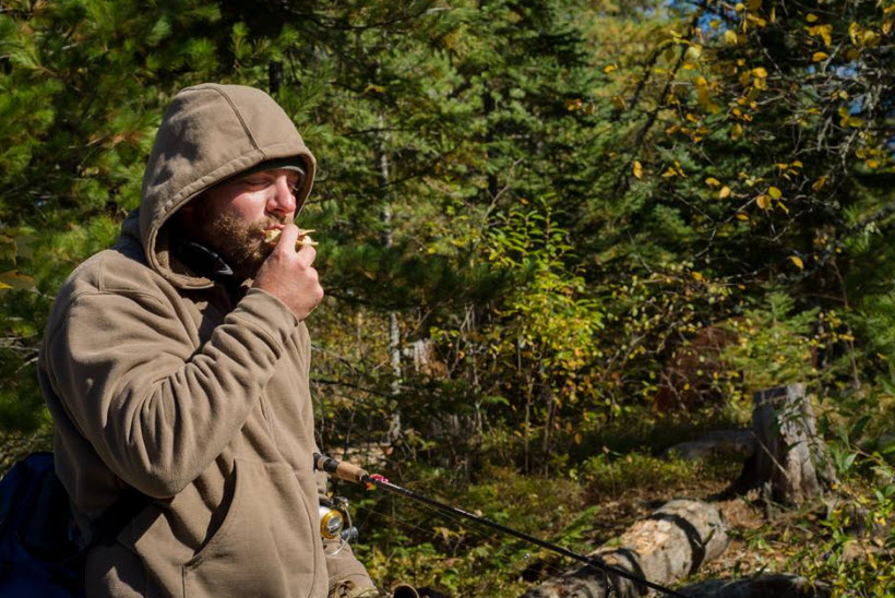 A person, wearing a hoodie, has a fishing pole under their arm, with the forest in the background