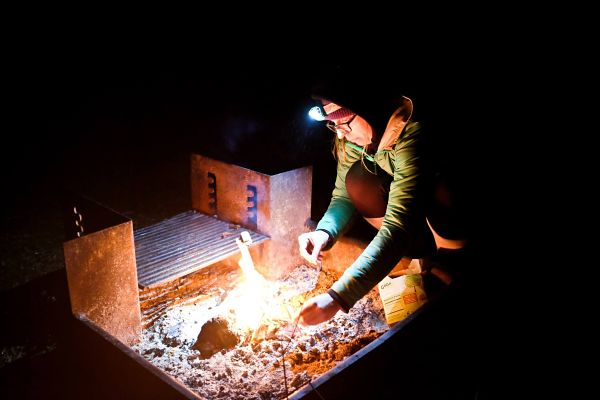 A person, bent over a fire in a camp stove on the ground, at nighttime