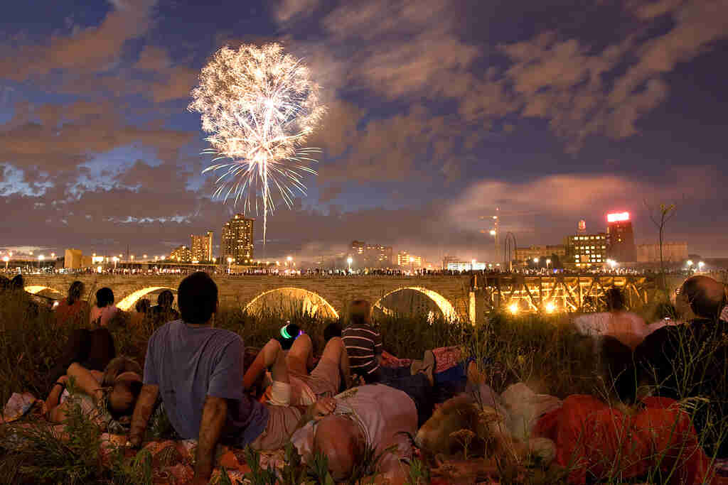 Rear view of people sitting in grass, watching fireworks, with a bridge and city skyline shown in front of them at night