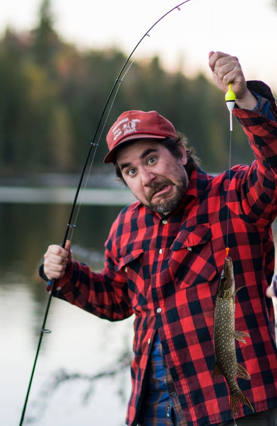Front view of a person making a funny face while holding up a fishing pole with a small northern pike fish on the line