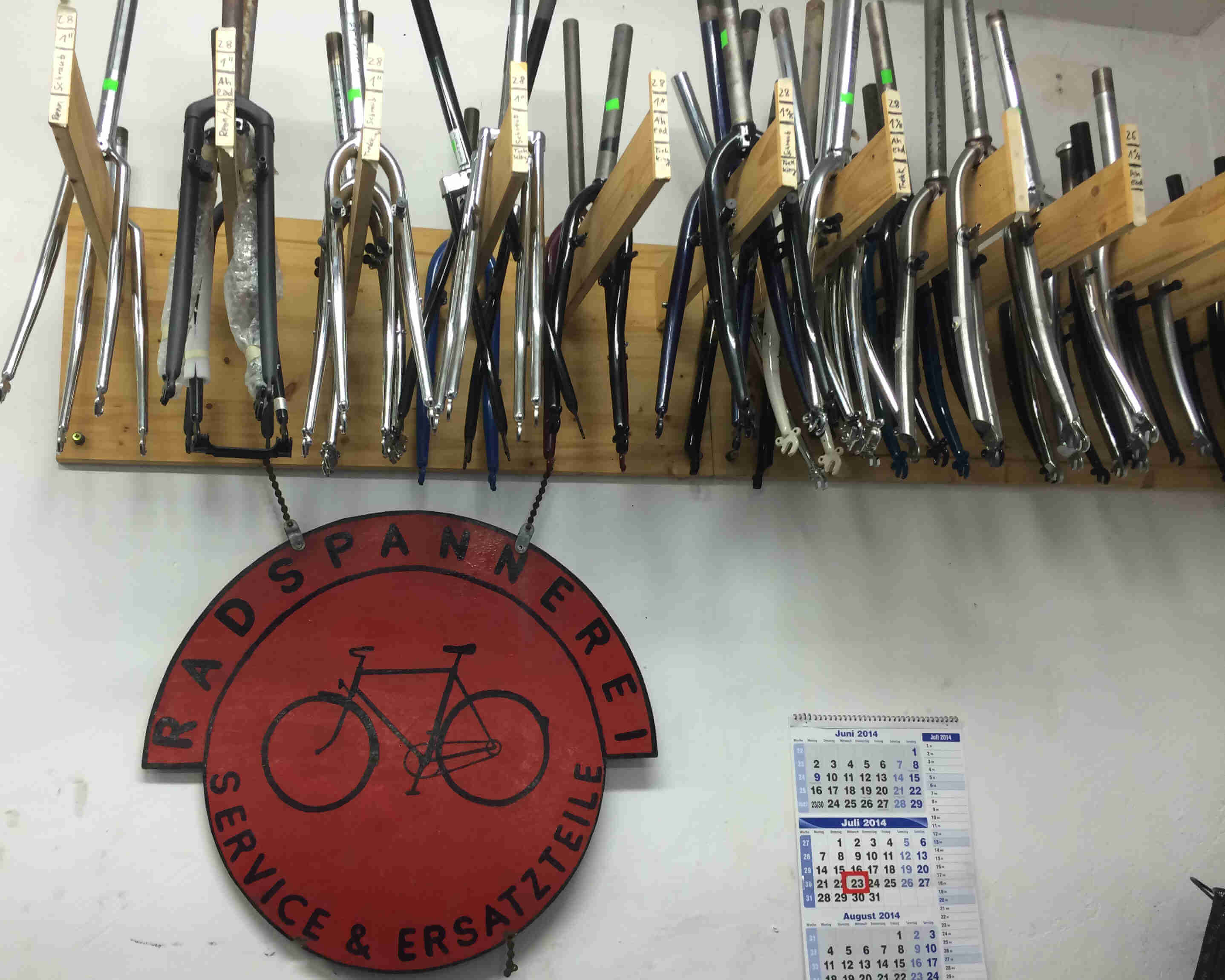 Upward, front view of a rack, full of bike forks, mounted to a wall with a Radspannerei sign below it