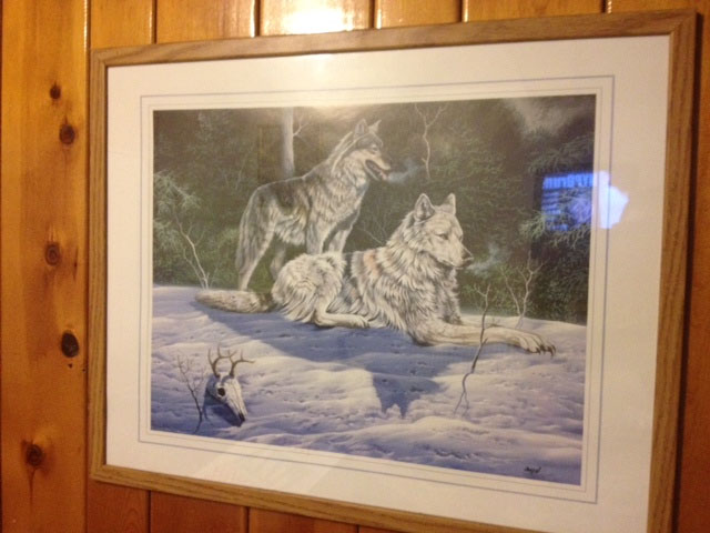 A framed, painted portrait, mounted to a wood wall, showing a wolf standing behind another that's laying down on snow