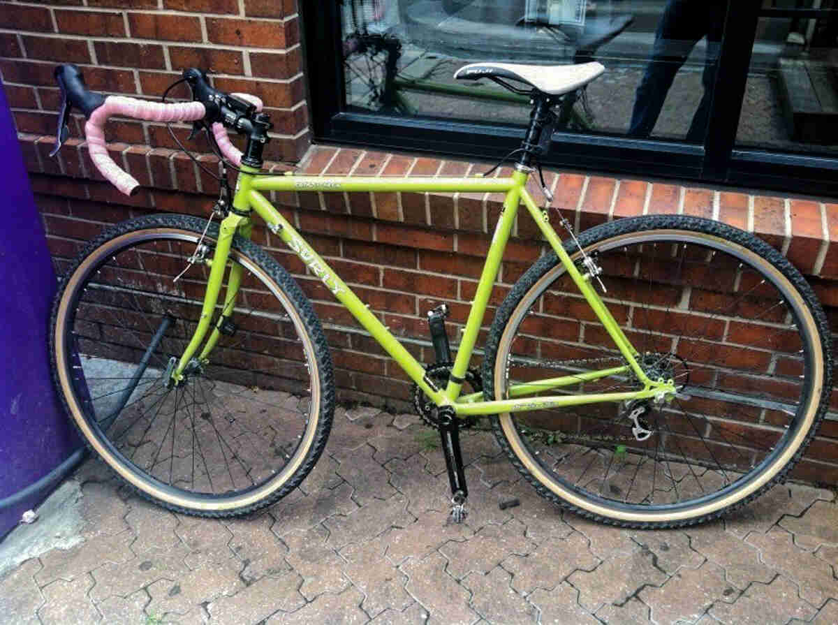 Left side view of a lime green Surly bike with pink handlebar, parked against a brick wall and window of a building