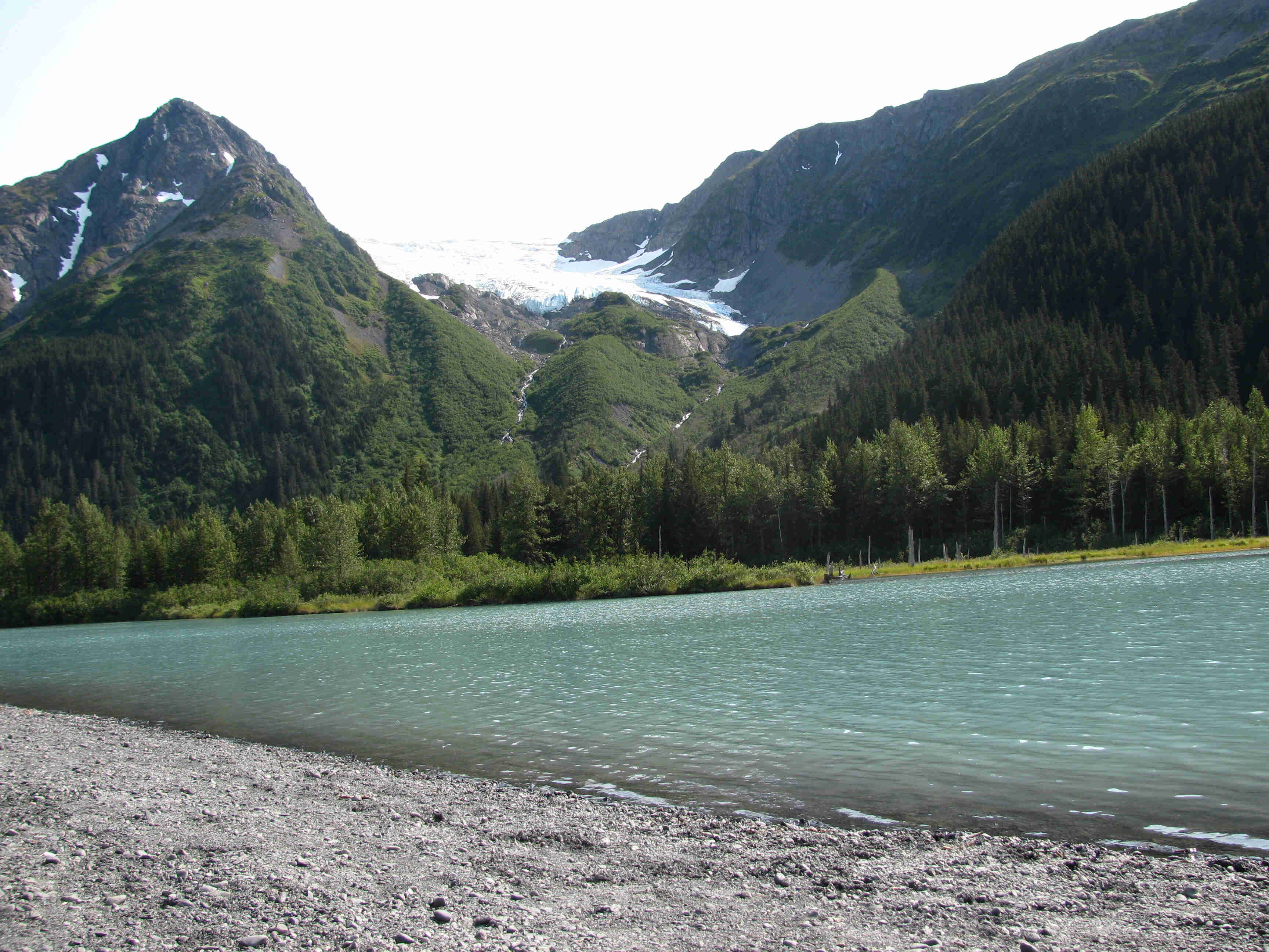 A turquoise colored lake, at the base of mountains with a glacier in a valley between them