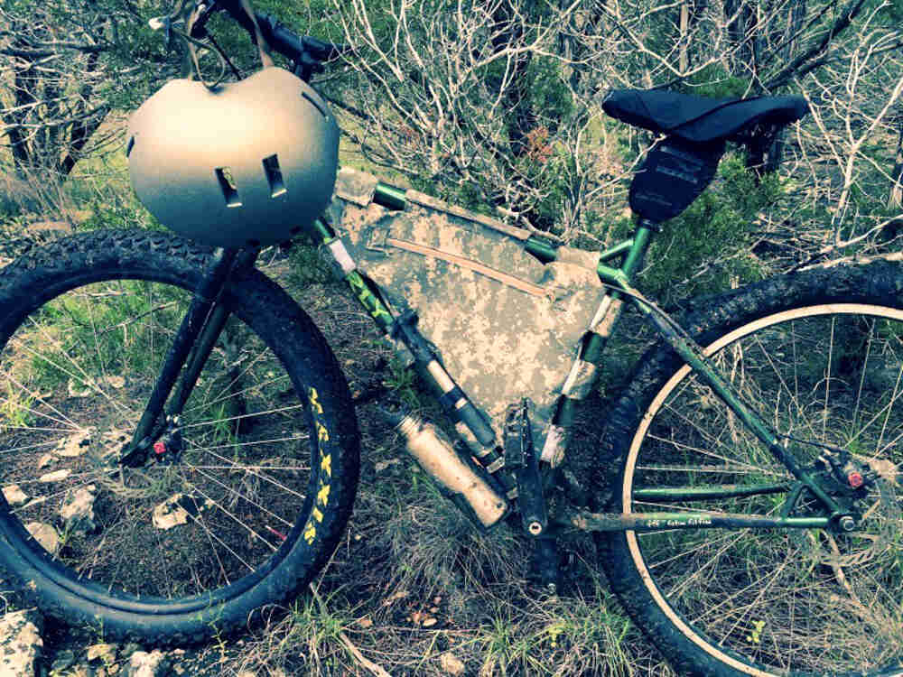 Left side view of a green Surly Krampus bike with a frame bag and helmet hanging from the handlebar, parked in brush