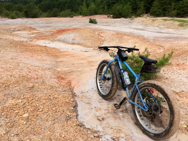 Rear, left side view of a light blue Surly fat bike, parked against a small bush in a gravel pit 