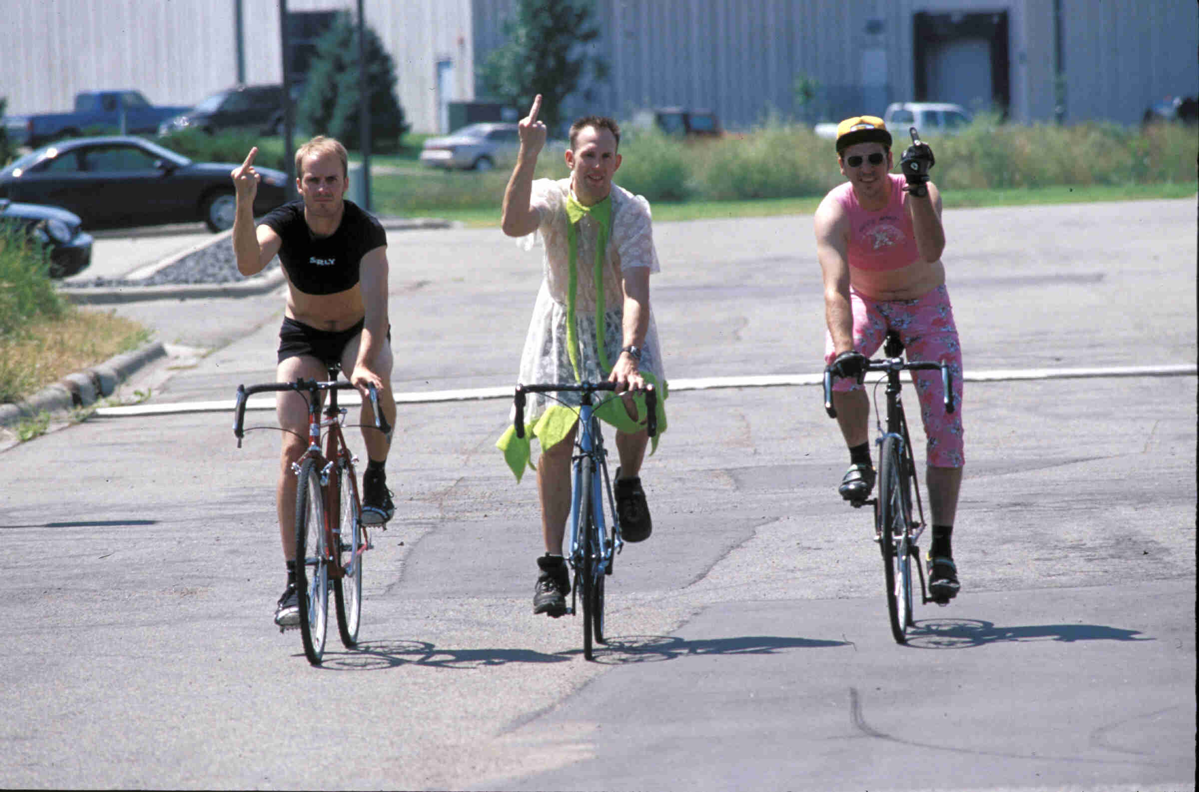 Front view of 3 cyclists wearing women's attire, holding up a middle finger as they ride their bikes on a paved street