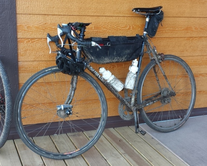 Left side view of a dirty, black Surly Pacer bike with gear, parked along a wood sided wall