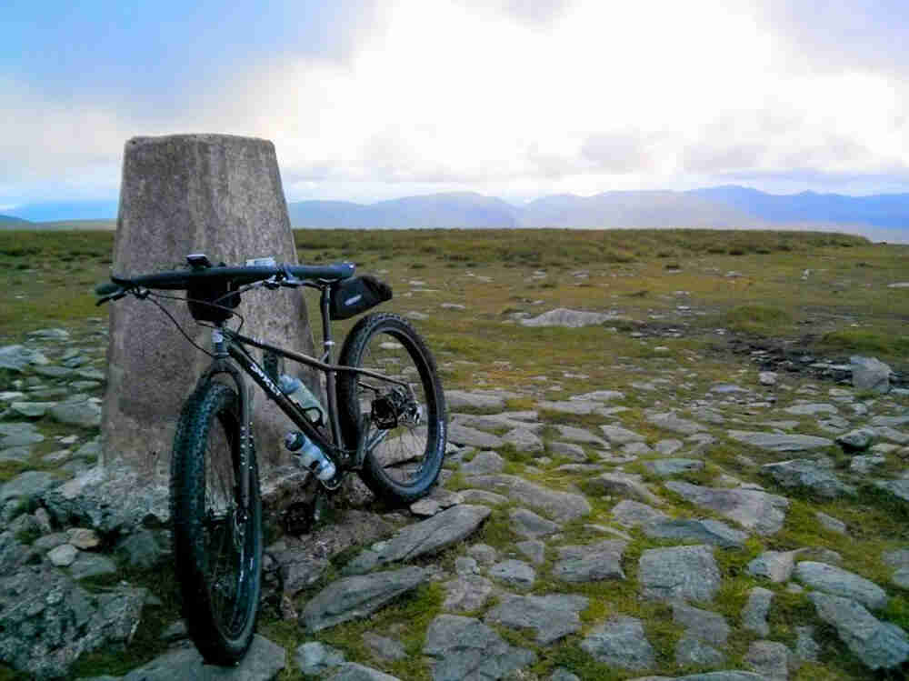 Front, left side view of a Surly bike, leaning on a stone block in a rocky grass field, with mountains in the background