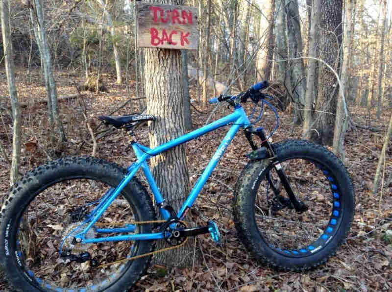 Right side view of a blue Surly Ice Cream Truck fat bike, in the wood, against a tree with a, Turn Back, sign on it