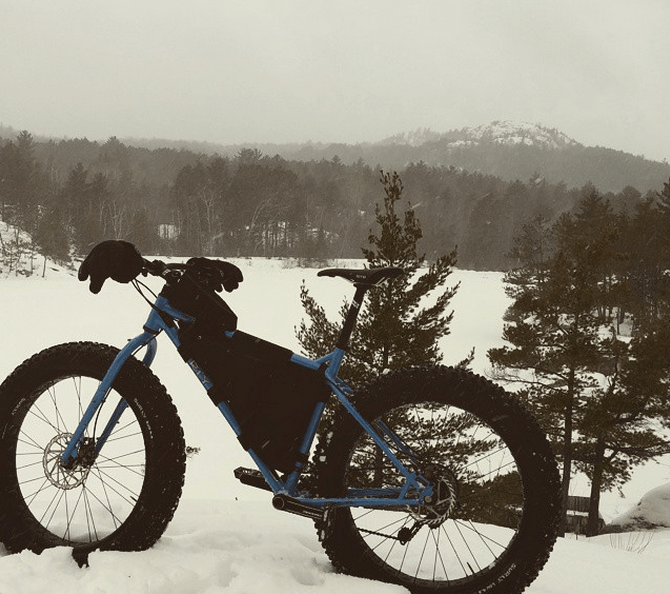 Left side view of a blue Surly Ice Cream Truck fat bike, in a snow covered field with trees and mountains in background