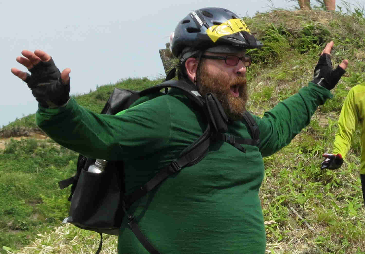 Front, right side, waist up view of a bearded person wearing a bike helmet, standing with their arms outstretched