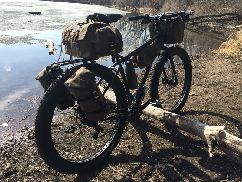 Rear, right side view of a black Surly bike with gear packs, parked on the dirt shore line of a thawing pond