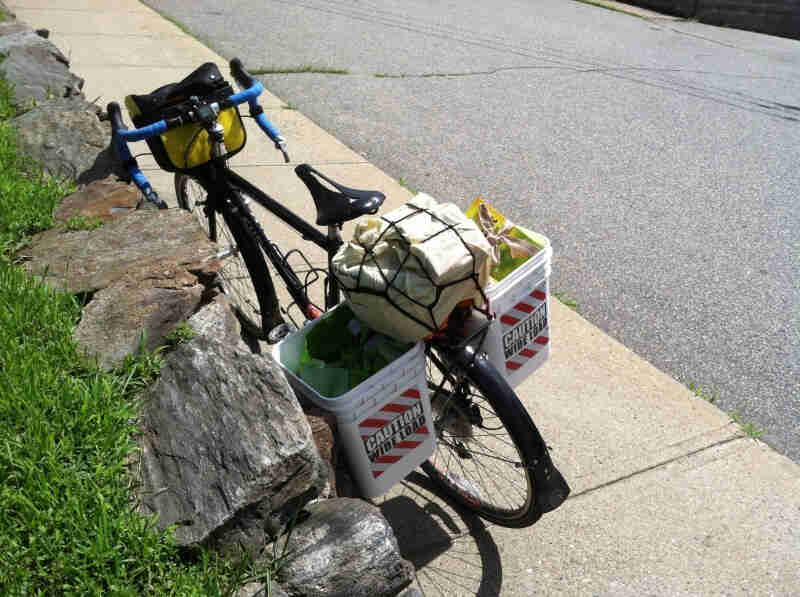 Downward, rear view of a Surly bike, with buckets on the sides of the rear rack, on a sidewalk against a stone wall