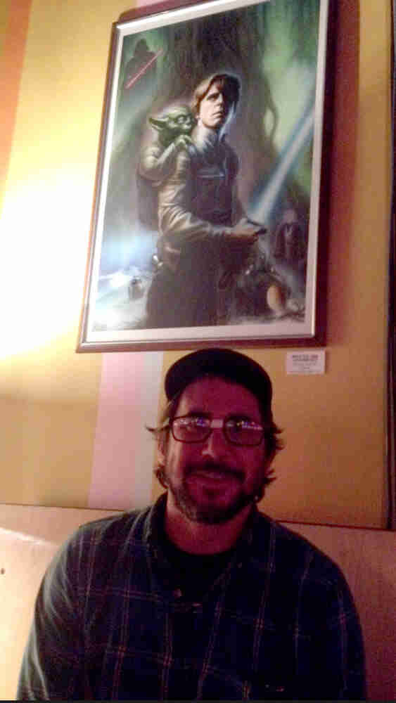 A person wearing glasses with tape on the bridge, sits under a painting of Luke Skywalker and Yoda, on wall behind