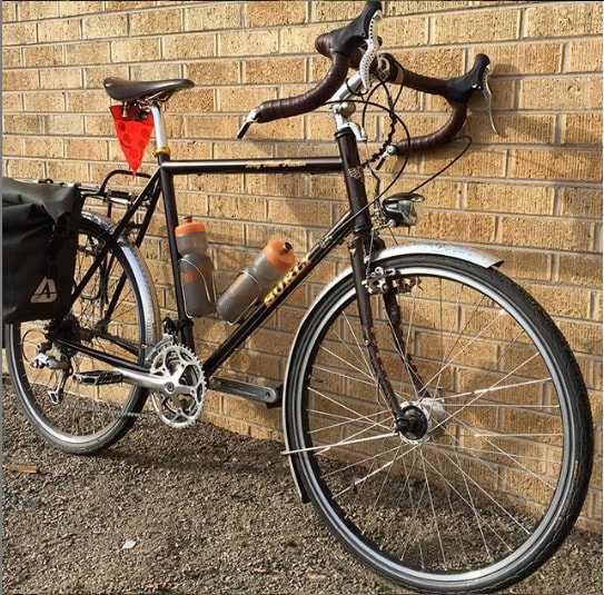 A black Surly road bike with fenders, saddle bags and two water bottles leans against a light brown brick wall 