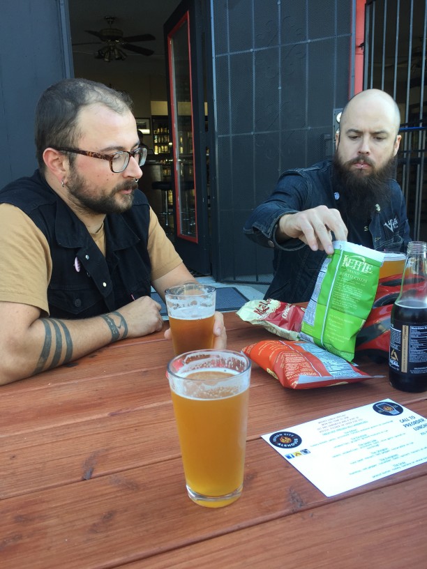 2 people sitting at a table, with glasses of beer and bags of potato chips