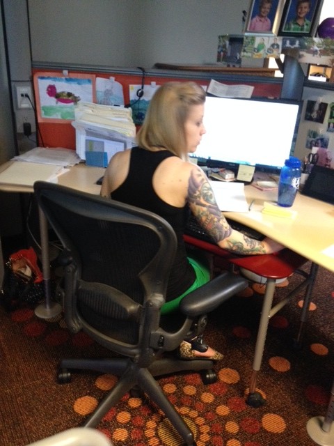 Rear view of a person with a tattooed right arm, sitting at a desk, in an office cubicle