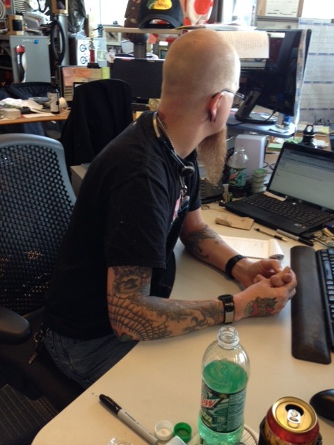 Right side view of a person, with a shaved head and tattooed arms, sitting at a desk, in an office cubicle
