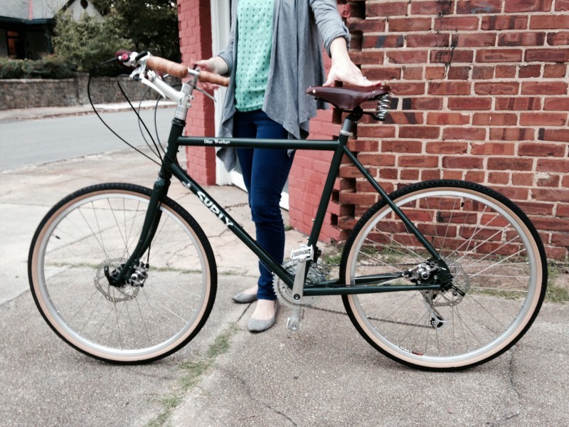 Left side view of a green Surly Disc Trucker bike with a cyclist standing behind, on a sidewalk