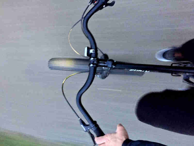 Downward, cropped view of the front of a black Surly bike, with a cyclist riding down a paved road