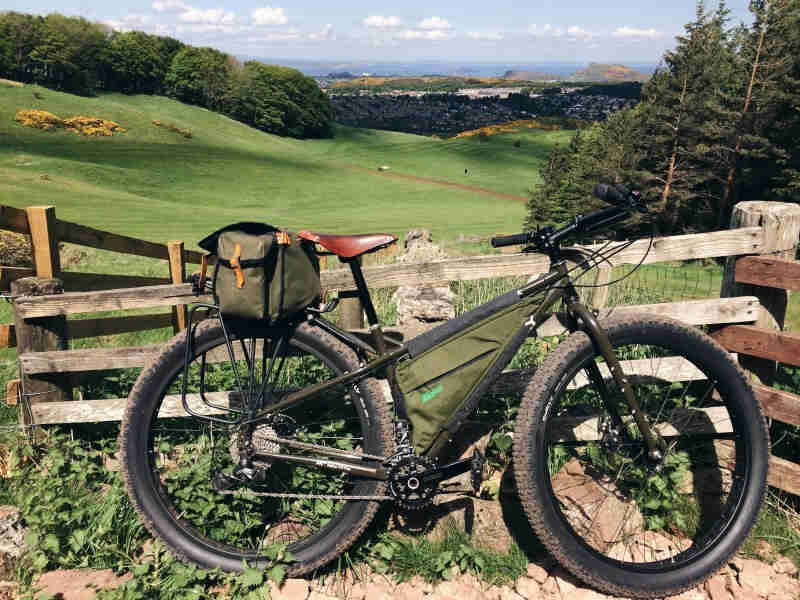 Right side view of an olive drab Surly fat bike, leaning on a wood gate, with a hilly grass field and trees behind it