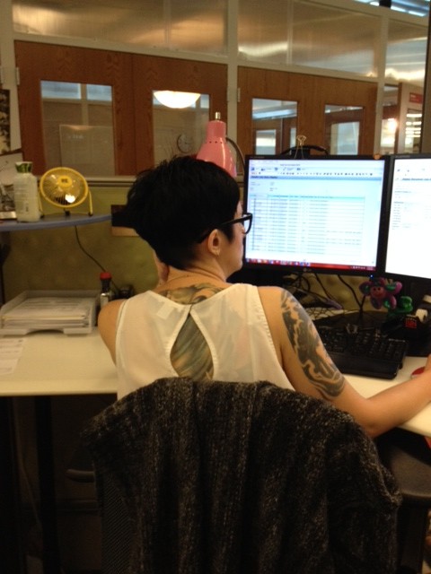 Right view of a person, with a tattooed back and right arm, sitting at a desk in an office cubicle