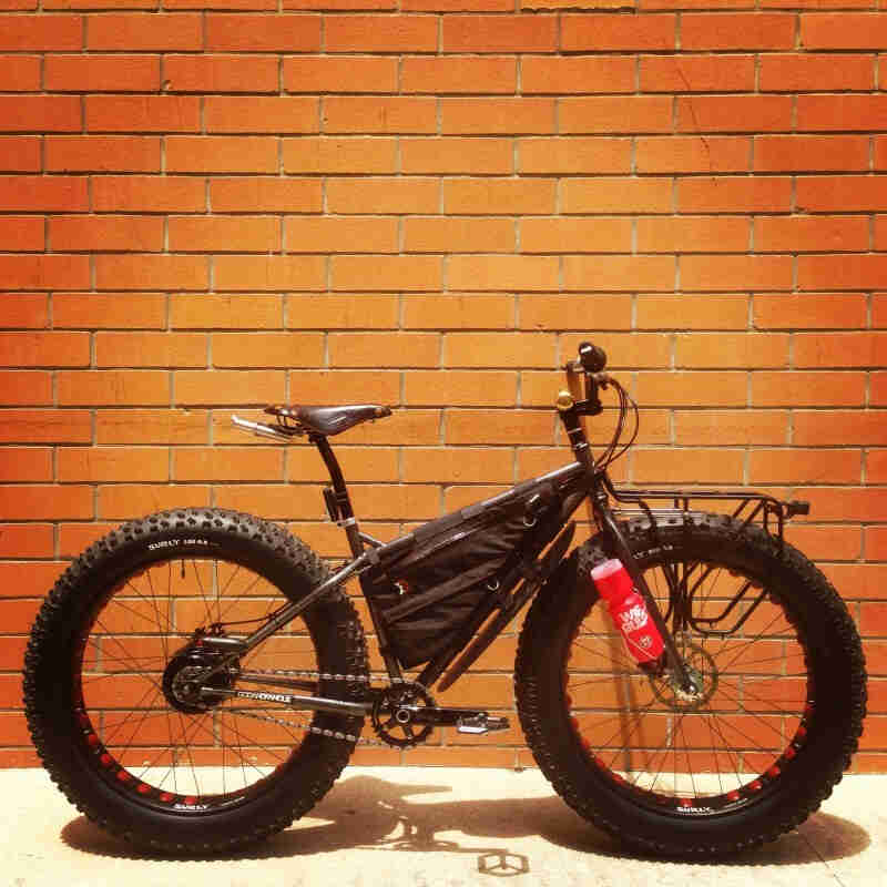 Right side view of a Surly fat bike with a frame bag, parked on a sidewalk, leaning on an orange brick wall